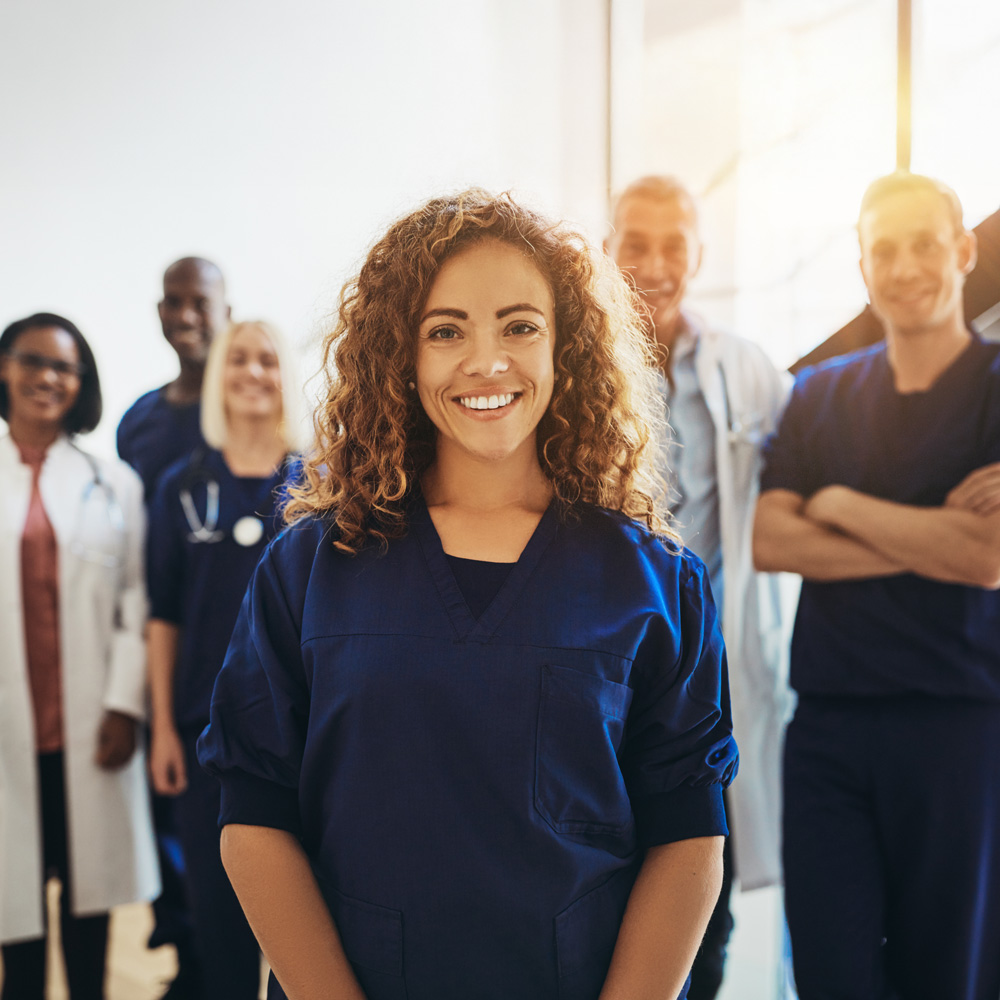 Im Vordergrund ist eine freundliche medizinische Fachangestellte zu sehen. Sie lächelt in die Kamera. Im Hintergrund steht ein Team aus Kolleginnen, Ärztinnen und Ärzten.